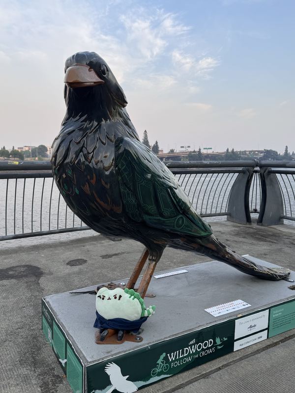 Pusheeno at the feet of a large painted crow sculpture beside a river