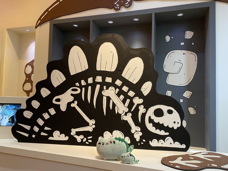 Pusheeno and a tiny dinosaur-cat plush in front of a cartoon stegosaurus skeleton mural