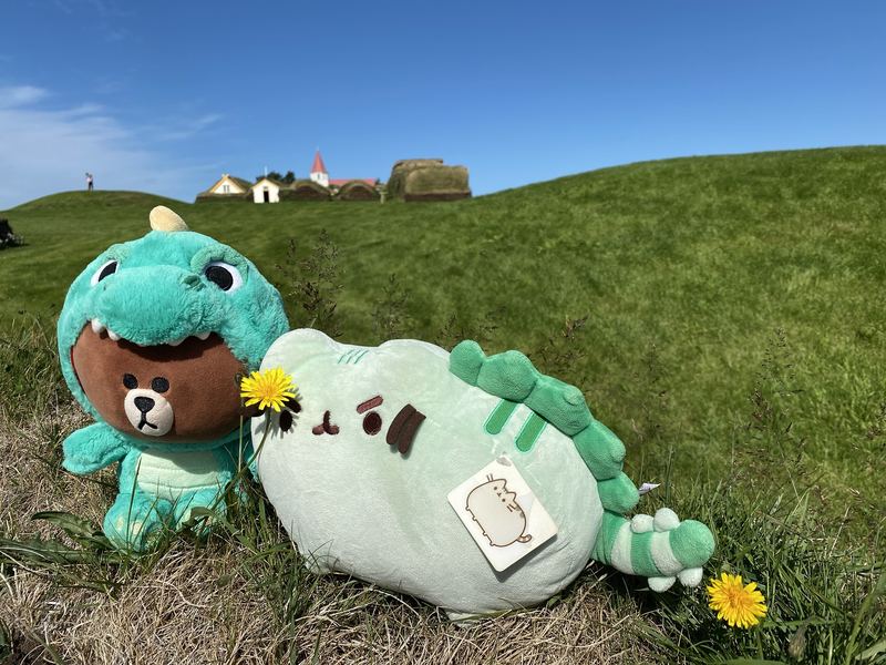 Pusheeno and Dino Brown, a bear in a teal dinosaur costume, on a grassy hill with turf houses and a small church behind