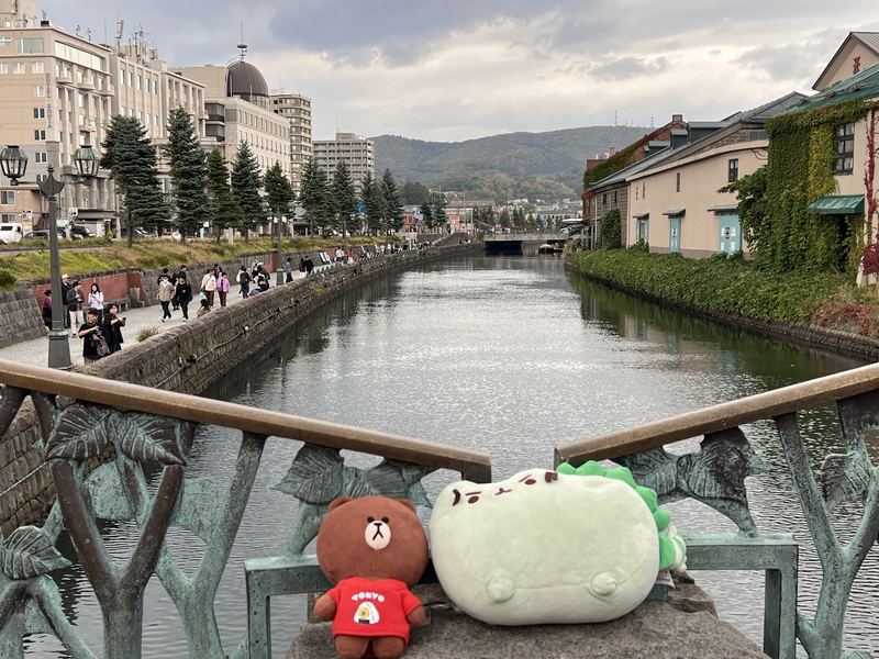 Pusheeno and Brown, a bear in a red Tokyo shirt, on a bridge railing above a canal lined with old stone warehouses