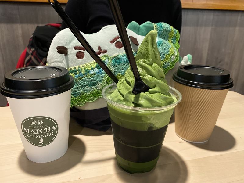 Pusheeno behind a tall matcha soft-serve float with two spoons, between two takeaway cups