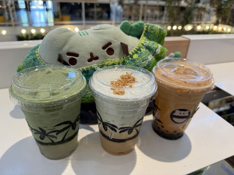 Pusheeno in his crocheted collar behind three takeaway drinks: a matcha frappe, a cream-topped milk tea and a Thai iced tea with boba