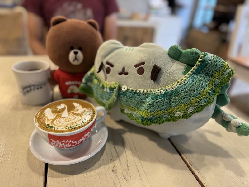 Brown, a bear in a red Tokyo shirt, and Pusheeno in his crocheted collar beside a latte with swan latte art dusted with matcha
