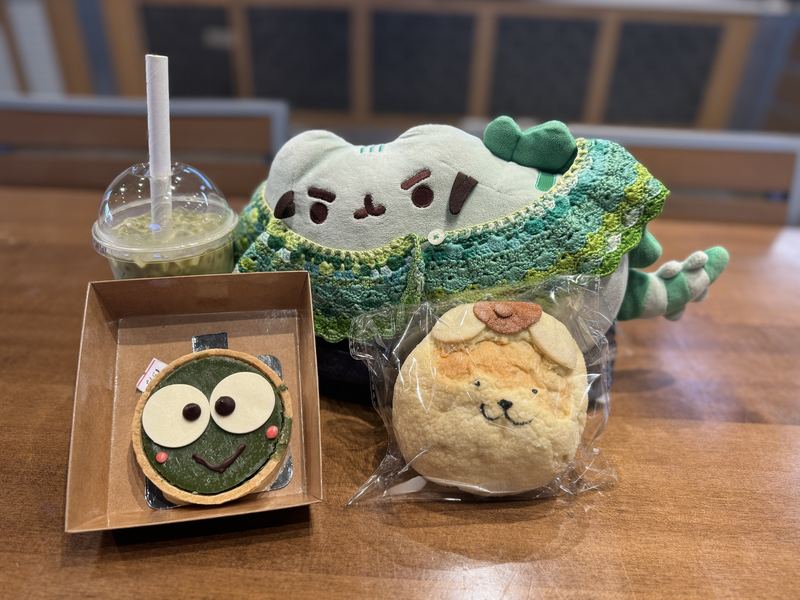 Pusheeno in his crocheted collar with an iced matcha, a green Keroppi-faced tart in a box and a bun shaped like Pompompurin