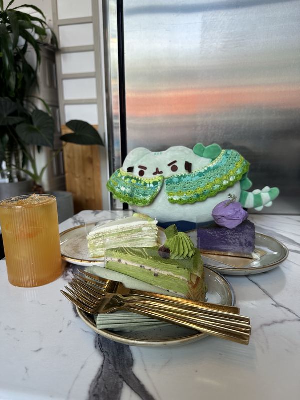Pusheeno in his crocheted collar behind three slices of cake: matcha crepe, pandan crepe and purple ube