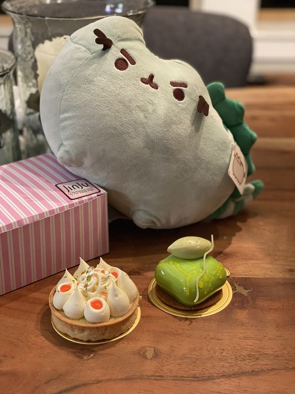 Pusheeno leaning on a pink striped JinJu Patisserie box, with a meringue-topped tart and a green glazed cake in front