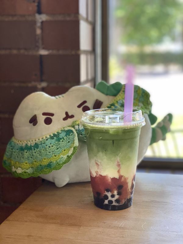 Pusheeno in his crocheted collar behind a layered matcha and strawberry boba drink with a purple straw