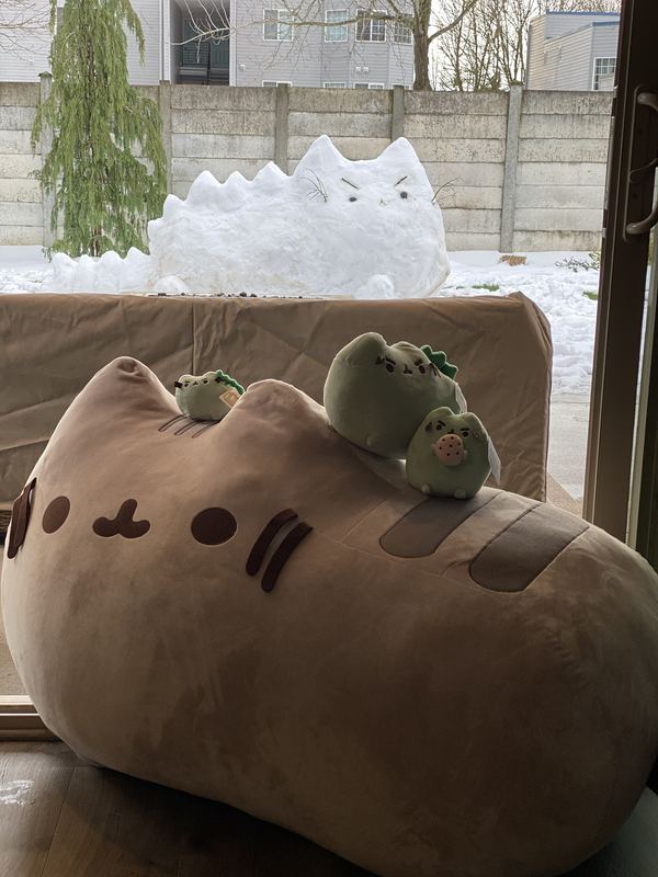 Pusheeno and two minis on top of a giant Pusheen plush by a window, with a large snow sculpture of a Pusheenosaurus in the snowy yard outside